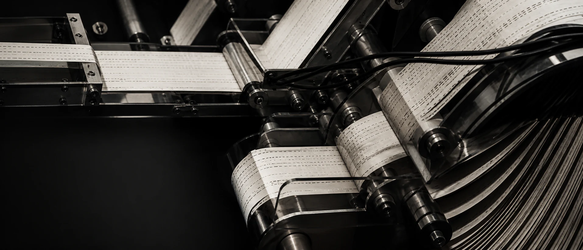 Punched paper tape threading through polished metal rollers and dark routing machinery.